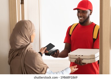 Fast Food Delivery And Cashless Contactless Payment. Smiling Black Male Courier Guy Wearing Red Uniform Holding Point Of Sale Terminal And Pizza Boxes, Muslim Woman In Hijab Paying Using Smartphone