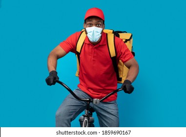 Fast Food Delivery. Portrait of black courier man wearing medical face mask, protective gloves and yellow thermo backpack, riding bicycle isolated on blue studio background. Shipment during pandemic - Powered by Shutterstock