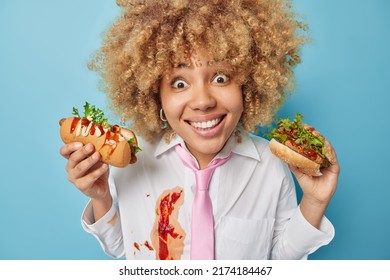 Fast Food To Ease Your Hunger. Positive Surprised Curly Haired Woman Eats Delicious Hambruger And Hot Dog Wears White Formal Shirt Dirty With Ketchup And Pink Tie Isolated Over Blue Background