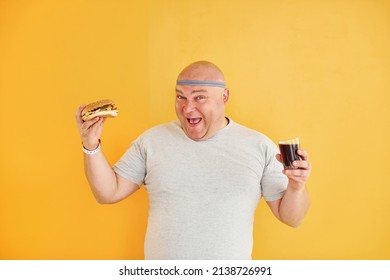 With Fast Food. Funny Overweight Man In Sportive Head Tie Is Against Yellow Background.