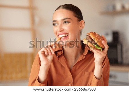 Fast food lover. Happy lady eating burger and french fries, biting chips and looking aside, sitting in kitchen at home. Playful woman holding sandwich and tasty potato