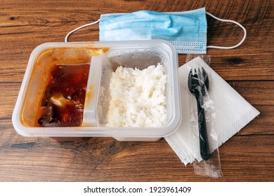 Fast Food Lunch On A Wooden Table With Blue Face Mask, Napkin, And Plastic Fork. Chinese Style Meal With White Rice And Bbq Chicken In A Plastic Container