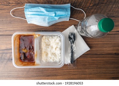 Fast Food Lunch On A Wooden Table With Blue Face Mask, Napkin, And Plastic Fork. Chinese Style Meal With White Rice And Bbq Chicken In A Plastic Container