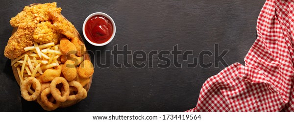 fast food meals : onion\
rings, french fries, chicken nuggets and fried chicken on dark\
table, top view