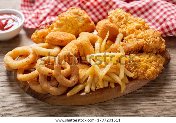 fast food meals : onion rings,\
french fries, chicken nuggets and fried chicken on a wooden\
table