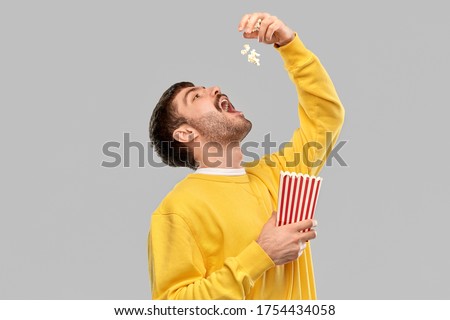 fast food people concept - young man in yellow sweatshirt eating popcorn throwing it to open mouth over grey background