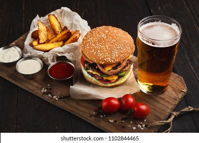 Fast Food Restaurant Dish. Juicy Burger, Potato Wedges, Sauces And Glass Of Cold Beer On Dark Wooden Background
