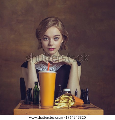 Fast food. Young woman in art action isolated on brown background. Retro style, comparison of eras concept. Beautiful female model like legendary chess player, queen or duchess, old-fashioned.