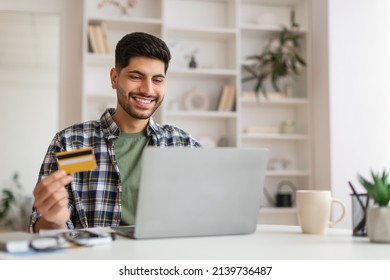 Fast Online Shopping. Portrait of smiling young man holding debit credit card in hand and using pc laptop, making financial transaction sitting at table in home office in living room, free copy space - Powered by Shutterstock