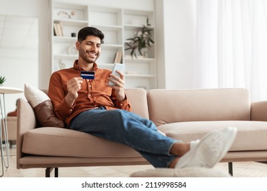 Fast Online Shopping. Smiling Young Arab Guy Holding Debit Credit Card In Hand And Using Cell Phone, Making Financial Transaction Sitting On The Couch At Home In Living Room, Free Copy Space