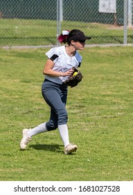 Fast Pitch Softball Outfielder Wearing Sunglasses Handling A Ball In Play On The Grass In The Outfield.