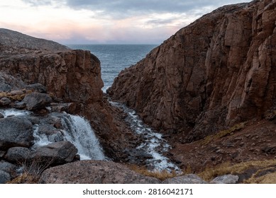 Fast river stream flowing through a rocky red granite canyon towards the cold arctic sea shore under a soft sunset sky.