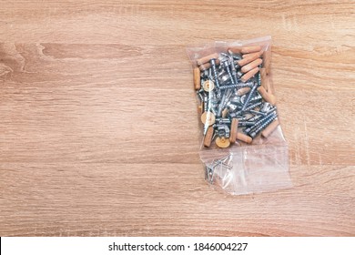 Fasteners Package For Assembling Furniture. Connecting Hardware, Screws, Eccentric Coupler, Wooden Dowel Pins, Nails And Screw Cover Caps In The Plastic Bag On Wooden Background With Copy Space.