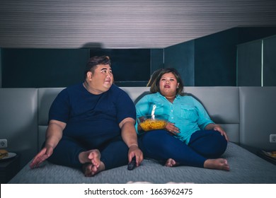 Fat Asian couple sitting on a bed, while eating instant fried noodle in their bedroom