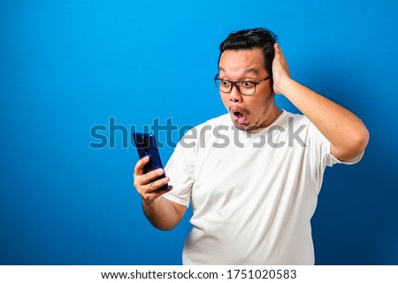 Fat Asian guy wearing a white t-shirt looks surprised at the good news he received from his smartphone. Men show shocked movements with bulging eyes while rolling down his glasses on a smartphone
