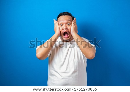 Fat Asian man in white t-shirt shows funny shocked and surprise expression against blue background