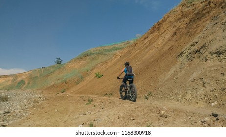 Fat Bike Also Called Fatbike Or Fat-tire Bike In Summer Driving On The Road. The Guy Rides By The Hill On A Sand Clay Path, Behind Him The Shore By The Sea.