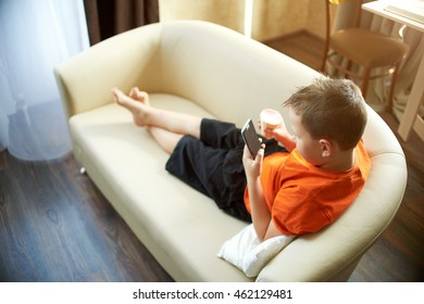 Fat Boy Is Lying On The Couch Playing With The Phone In The Room In An Orange Shirt Is Eating Ice Cream