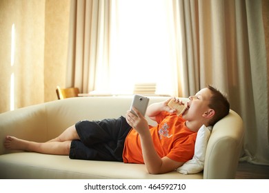 Fat Boy Is Lying On The Couch Playing With The Phone In The Room In An Orange Shirt Is Eating Ice Cream