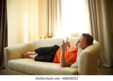 Fat Boy Is Lying On The Couch Playing With The Phone In The Room In An Orange Shirt Is Eating Ice Cream