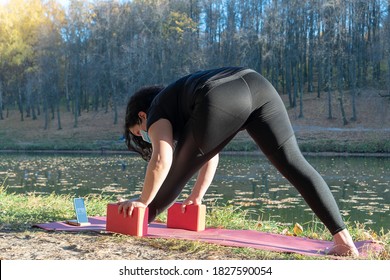 Fat Caucasian Woman With Long Curly Dark Hair Dressed In Black Tshirt And Black Sportive Pants In A Medical Mask Online Is Doing Yoga  In The Autumn Park. The Phone Shows An Online Yoga Class