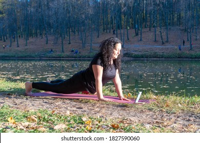 Fat Caucasian Woman With Long Curly Dark Hair Dressed In Black Tshirt And Black Sportive Pants Online Is Doing Yoga On Purple Mat Near The Lake In The Autumn Park. The Phone Shows An Online Yoga Class