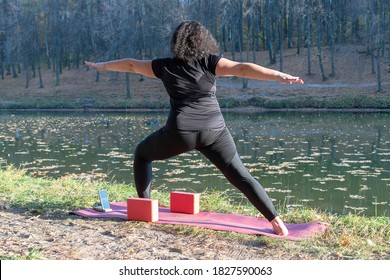 Fat Caucasian Woman With Long Curly Dark Hair Dressed In Black Tshirt And Black Sportive Pants Online Is Doing Yoga On Purple Mat Near The Lake In The Autumn Park. The Phone Shows An Online Yoga Class