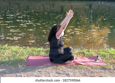 Fat Caucasian Woman With Long Curly Dark Hair Dressed In Black Tshirt And Black Sportive Pants In A Medical Mask Online Is Doing Yoga  In The Autumn Park. The Phone Shows An Online Yoga Class