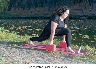 Fat Caucasian Woman With Long Curly Dark Hair Dressed In Black Tshirt And Black Sportive Pants Online Is Doing Yoga On Purple Mat Near The Lake In The Autumn Park. The Phone Shows An Online Yoga Class