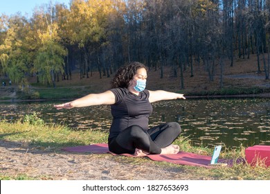 Fat Caucasian Woman With Long Curly Dark Hair Dressed In Black Tshirt And Black Sportive Pants In A Medical Mask Online Is Doing Yoga  In The Autumn Park. The Phone Shows An Online Yoga Class