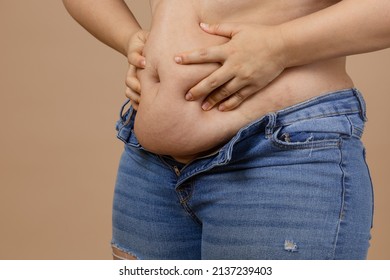 Fat female adult showing fat belly pressing on it with hands wearing blue jeans on beige background. Sudden weight gain. Visceral fat. Body positive. Tight little clothes. Need for wardrobe change.