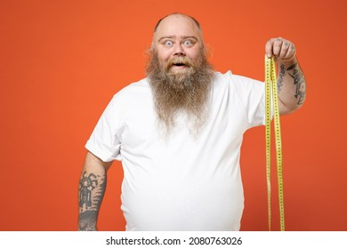 Fat fun smiling caucasian pudge obese chubby overweight tattooed bearded big belly man 30s wearing white t-shirt holding meter measure tape isolated on red orange color background studio portrait.