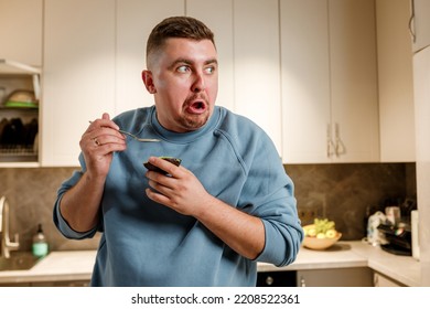 Fat And Funny Man Eating Avocado While Standing In Modern Home Kitchen. Slimming And Healthy Lifestyle Concept