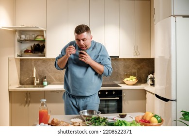 Fat And Funny Man Eating Avocado While Standing In Modern Home Kitchen. Slimming And Healthy Lifestyle Concept