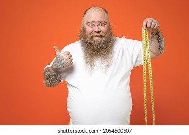 Fat happy pudge obese chubby overweight tattooed bearded big belly man 30s wearing white t-shirt holding meter measure tape show thumb up like gesture isolated on orange background studio portrait.