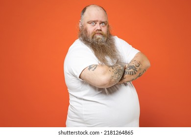 Fat indignant pudge obese chubby overweight tattooed blue-eyed bearded man 30s has big belly wearing white t-shirt holding hands crossed folded isolated on red orange color background studio portrait