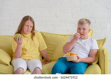 Fat Kids Eating On Couch. Girl Is Eating An Apple And Jealous Because Of Boy Eating Chips. Diet Challenge.