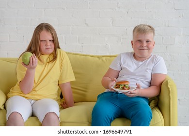 Fat Kids Eating On A Couch. Girl Is Jealous Of Boy Eating Tasty Hamburger While She's Having An Apple.