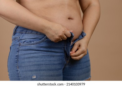 Fat lady with flabby belly with hands in process of zipping up jeans on beige background. Sudden weight gain. Visceral fat. Body positive. Tight little clothes. Need for wardrobe change.