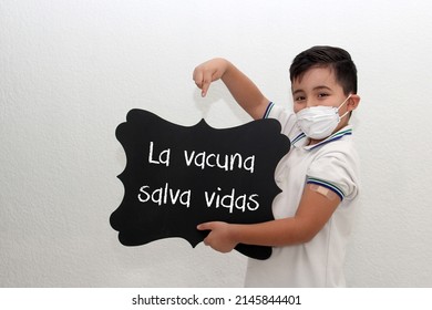 Fat Little Hispanic Boy With Face Mask And School Uniform Shirt Shows His Recently Vaccinated Arm Against Covid-19 With A Black Sign That Says 