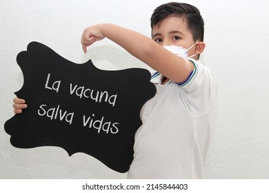 Fat Little Hispanic Boy With Face Mask And School Uniform Shirt Shows His Recently Vaccinated Arm Against Covid-19 With A Black Sign That Says 