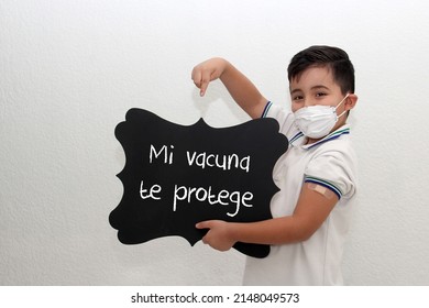 Fat Little Hispanic Boy With Face Mask And School Uniform Shirt Shows His Recently Vaccinated Arm Against Covid-19 With A Black Sign That Says 