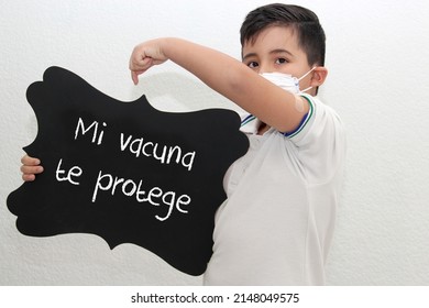 Fat Little Hispanic Boy With Face Mask And School Uniform Shirt Shows His Recently Vaccinated Arm Against Covid-19 With A Black Sign That Says 