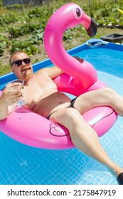 A Fat Man With A Beard And Mustache In Sunglasses With Alcohol Martini Glass And Smokes A Cigar Bathes In A Framed Pool With  Water And A Pink Inflatable Flamingo Under A Blue Sky In A Poor Area