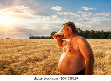 A fat man drinking beer in the field of barley and the sun goes to the sunset. A glass of beer is held by a fat man in hand. A large glass of light beer.