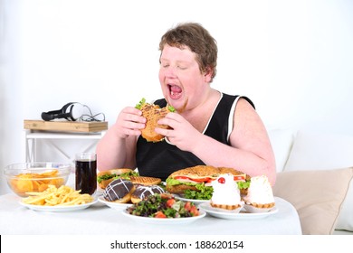 Fat man eating a lot of unhealthy food, on home interior background  