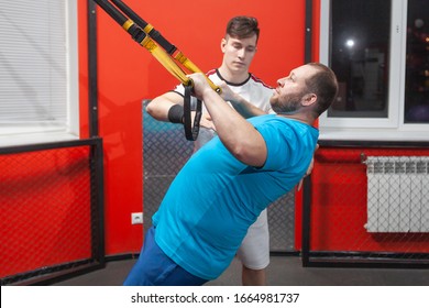 Fat Man In A Gym Is Diligently Doing Pull-ups Under The Guidance Of A Personal Trainer. Overweight TRX