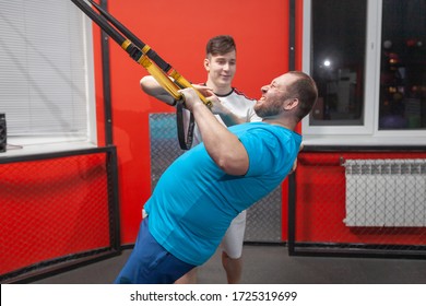 Fat Man In A Gym Is Diligently Doing Pull-ups Under The Guidance Of A Personal Trainer. Overweight TRX