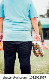 Fat Man Holding Football Boots And Stands Watching Soccer Practice.