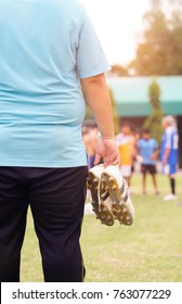 Fat Man Holding Football Boots And Stands Watching Soccer Practice.